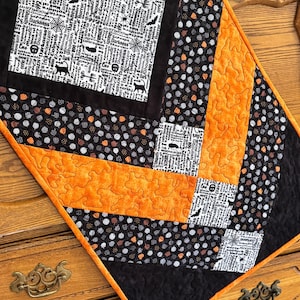 May include: A quilted table runner featuring a chevron pattern with alternating sections of black fabric with white Halloween-themed text, black fabric with orange and white polka dots, and solid orange fabric with a textured, swirling stitch pattern.