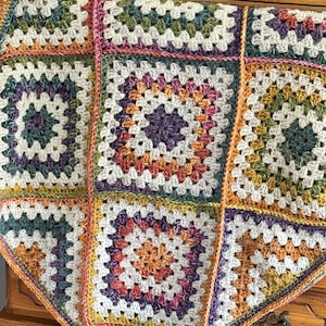 May include: A colourful, handmade granny square blanket. The blanket is made from yarn and features a patchwork design with squares in shades of white, green, purple, orange, and yellow. It has a cream-coloured border and a textured appearance.