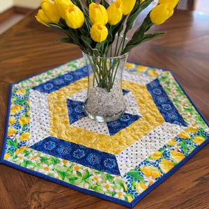May include: A hexagonal table runner with a lemon and floral pattern in shades of yellow, blue, and green. The runner is quilted and has a blue border. A vase with yellow tulips is on top of the runner.