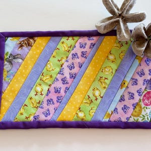 May include: A purple-trimmed quilted coaster with a diagonal pattern of yellow, green, purple, and pink fabric. The fabric features a variety of Easter-themed designs, including chicks, butterflies, and bunnies.