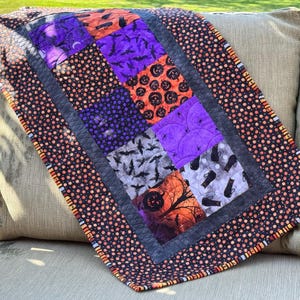 May include: A rectangular quilted table runner featuring a patchwork design of Halloween-themed fabrics. The centre squares display orange pumpkins, purple bats, black cats, and spider webs, framed by a black border with orange and white polka dots.