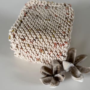 May include: A stack of square, hand-knitted coasters made from cream-coloured yarn with small, multicoloured flecks of orange, brown, and yellow throughout. Two dried, star-shaped botanical seed pods are positioned in front of the stack.
