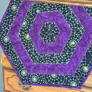 Peut inclure: Une nappe hexagonale sur le thème d'Halloween. Le centre et les bordures sont violets, avec un motif de toile d'araignée noire. Les bords sont cousus avec un fil contrastant, et le design est symétrique.