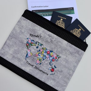 May include: A gray travel document holder with a black zipper and a floral world map design. The text "Rhonda's Travel Documents" is embroidered on the front. Two Canadian passports are visible inside the holder.