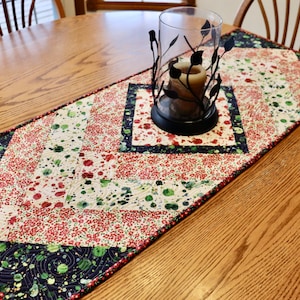 May include: A red, green and white patterned table runner with a black border. The runner is on a wooden table with a candle holder in the centre.