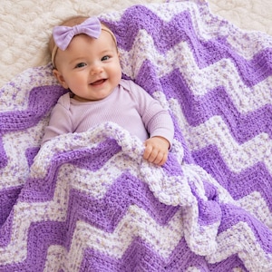 May include: A baby wearing a light purple long-sleeve ribbed shirt and a matching light purple bow headband, wrapped in a soft, textured crochet blanket featuring a repeating chevron pattern in alternating shades of purple and white.