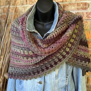 May include: A handmade crochet shawl in shades of brown, purple, and green. The shawl is draped over a denim jacket on a mannequin. The crochet pattern features a textured design with a scalloped edge, perfect for adding warmth and style.