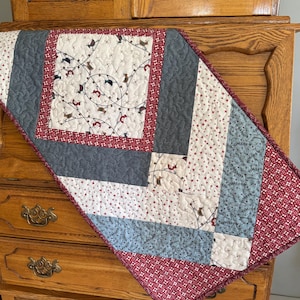 May include: A quilted table runner with a patchwork design. The runner features a central square with a pattern of string lights and small figures, surrounded by a red border. The design incorporates gray, white, and blue fabrics with star patterns.