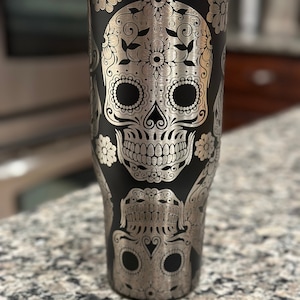 Puede incluir: Un vaso de acero inoxidable negro con un diseño de calavera de azúcar plateada. La calavera está rodeada de patrones florales.