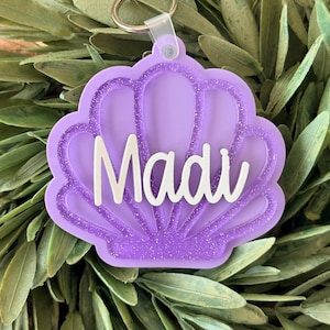 May include: A purple seashell-shaped keyring featuring a glittery finish and the name Madi in white script lettering. The keyring includes a clear plastic connector attached to a silver metal split ring for keys or bag attachment.