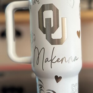 May include: White stainless steel travel mug with a black and silver OU logo, the name Makenna, and hearts. The mug has a black handle.
