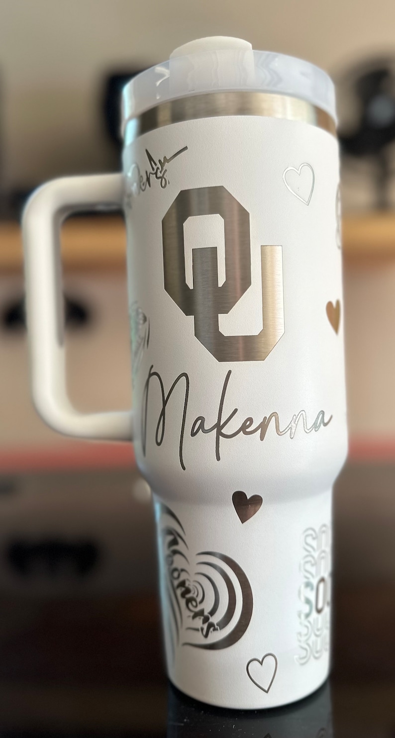 May include: White stainless steel travel mug with a black and silver OU logo, the name Makenna, and hearts. The mug has a black handle.