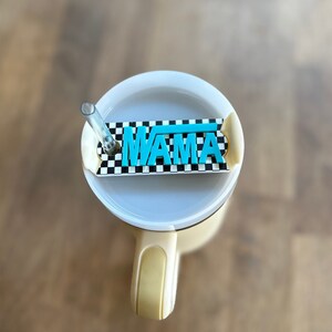 Puede incluir: Una taza de viaje beige con una tapa a cuadros en blanco y negro. La tapa tiene una inserción de plástico azul con la palabra "MAMA" escrita en ella.