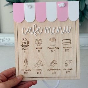 May include: A wooden cafe menu sign featuring a pink and white striped awning top. The board displays the text cafe menu and lists items including coffee, pretzel, french fries, macaron, ice cream, pizza, cinnamon bun, and croissant with prices.