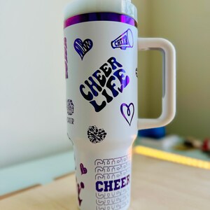 May include: White stainless steel travel mug with a purple and white design. The mug has a handle and features the text "Cheer Life" and other cheer-related graphics.