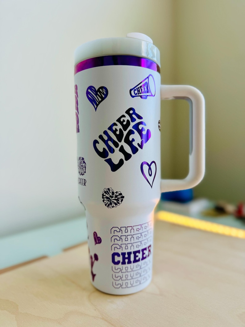 May include: White stainless steel travel mug with a purple and white design. The mug has a handle and features the text "Cheer Life" and other cheer-related graphics.