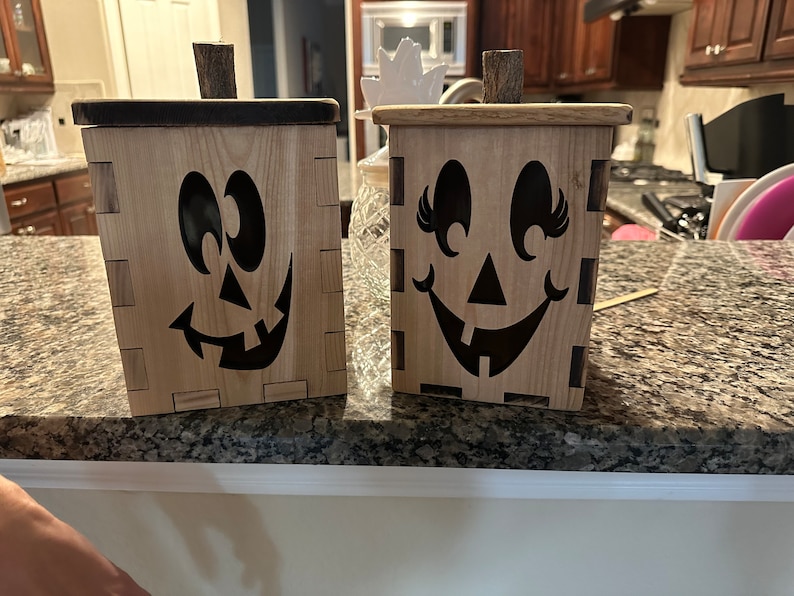 Puede incluir: Dos cajas de madera de calabaza de Halloween con caras pintadas de negro. Una caja tiene una cara sonriente con pesta&ntilde;as y la otra tiene una cara de calabaza de Halloween m&aacute;s tradicional.
