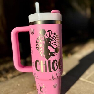 May include: Pink stainless steel tumbler with a gold handle and lid. The tumbler has a gold design of a cheerleader with pom-poms and the name "CHLOE" on the side. The tumbler is decorated with hearts and a heartbeat symbol.