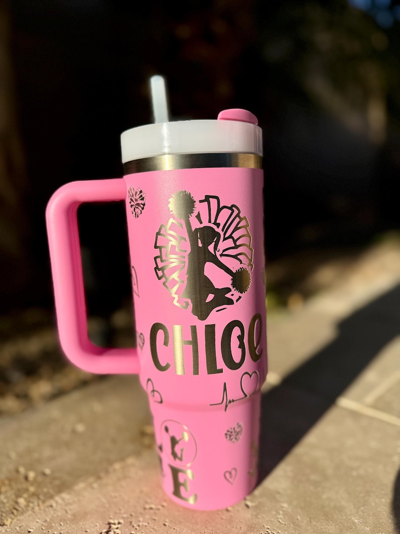 May include: Pink stainless steel tumbler with a gold handle and lid. The tumbler has a gold design of a cheerleader with pom-poms and the name "CHLOE" on the side. The tumbler is decorated with hearts and a heartbeat symbol.