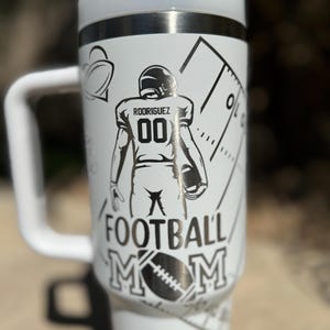 May include: White stainless steel travel mug with a black and white design. The design features a football player with the name "Rodriguez" and the number "00" on the back of the jersey. The text "Football Mom" is printed below the player.