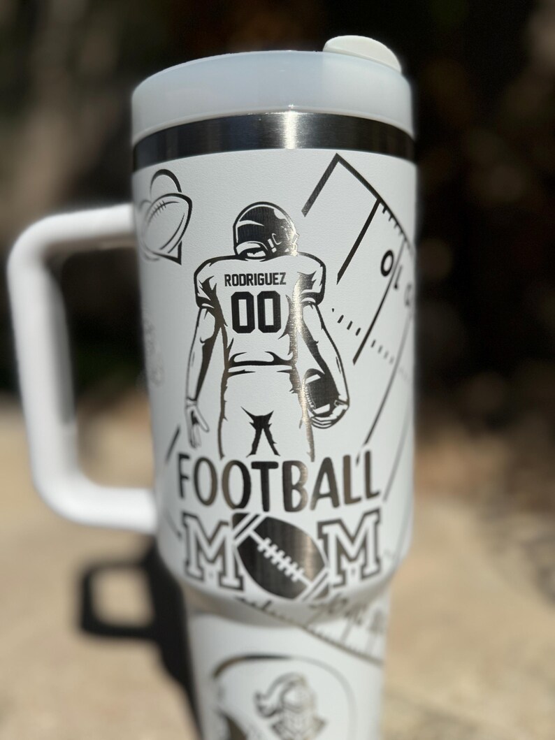 May include: White stainless steel travel mug with a black and white design. The design features a football player with the name "Rodriguez" and the number "00" on the back of the jersey. The text "Football Mom" is printed below the player.