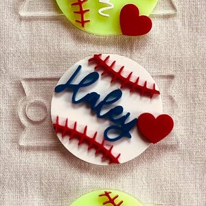 May include: Three softball-shaped ornaments with red stitching and a red heart. The first ornament is green and says "Taugau" in white. The second ornament is white and says "Haley" in blue. The third ornament is green and says "#32" in white.