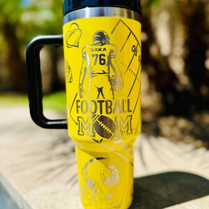 May include: Yellow stainless steel travel mug with a black handle and lid. The mug has a design of a football player with the number 76 and the words "Saka Football Mom".