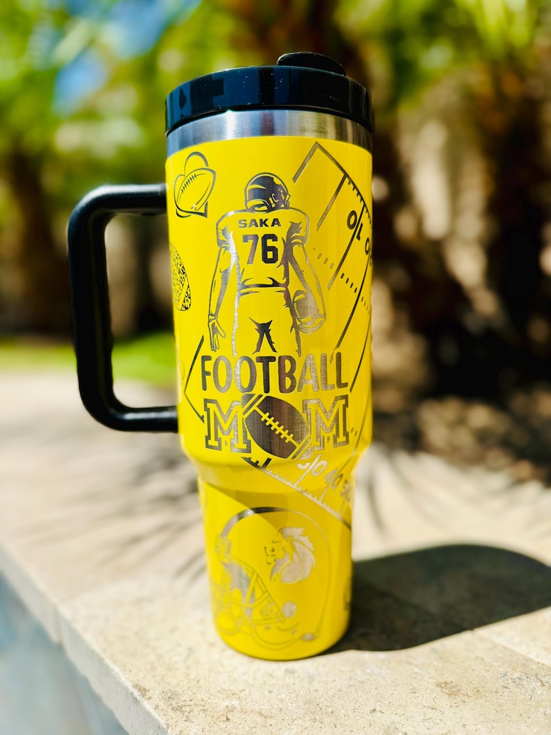 May include: Yellow stainless steel travel mug with a black handle and lid. The mug has a design of a football player with the number 76 and the words "Saka Football Mom".
