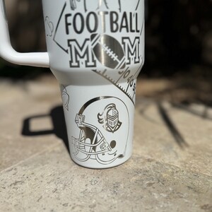 May include: White stainless steel tumbler with a black and white design. The design includes a football player, a football helmet, and the words "Football Mom".