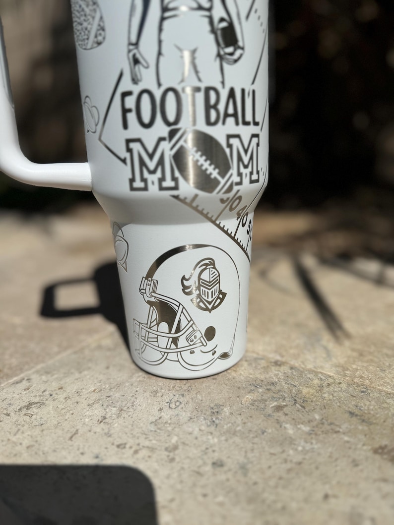 May include: White stainless steel tumbler with a black and white design. The design includes a football player, a football helmet, and the words "Football Mom".