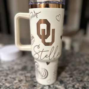 May include: White tumbler with a rose gold lid and a rose gold OU logo. The tumbler has a white handle and the name Stella is written on the side.