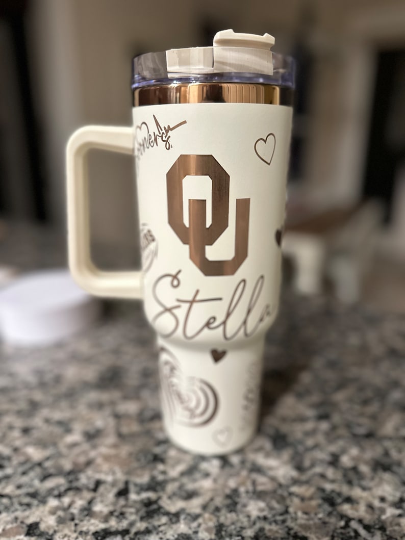 May include: White tumbler with a rose gold lid and a rose gold OU logo. The tumbler has a white handle and the name Stella is written on the side.