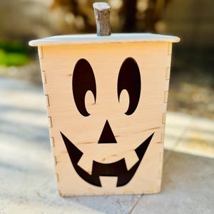Pode incluir: Uma caixa de madeira de abóbora de Halloween com um rosto esculpido e um pequeno pau de madeira para um caule. A caixa é de cor castanho claro e tem um rosto pintado de preto.