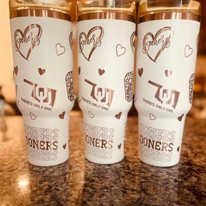 May include: Three white tumblers with a rose gold design. The tumblers feature a heart with the word "Sooners" inside, a heart with a stylized "OU" inside, and the text "There's only one." The tumblers also feature a repeating pattern of the word "Sooners" in a retro font.