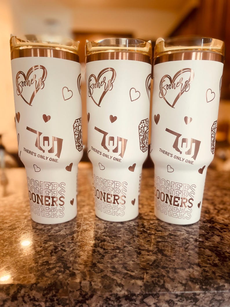 May include: Three white tumblers with a rose gold design. The tumblers feature a heart with the word "Sooners" inside, a heart with a stylized "OU" inside, and the text "There's only one." The tumblers also feature a repeating pattern of the word "Sooners" in a retro font.