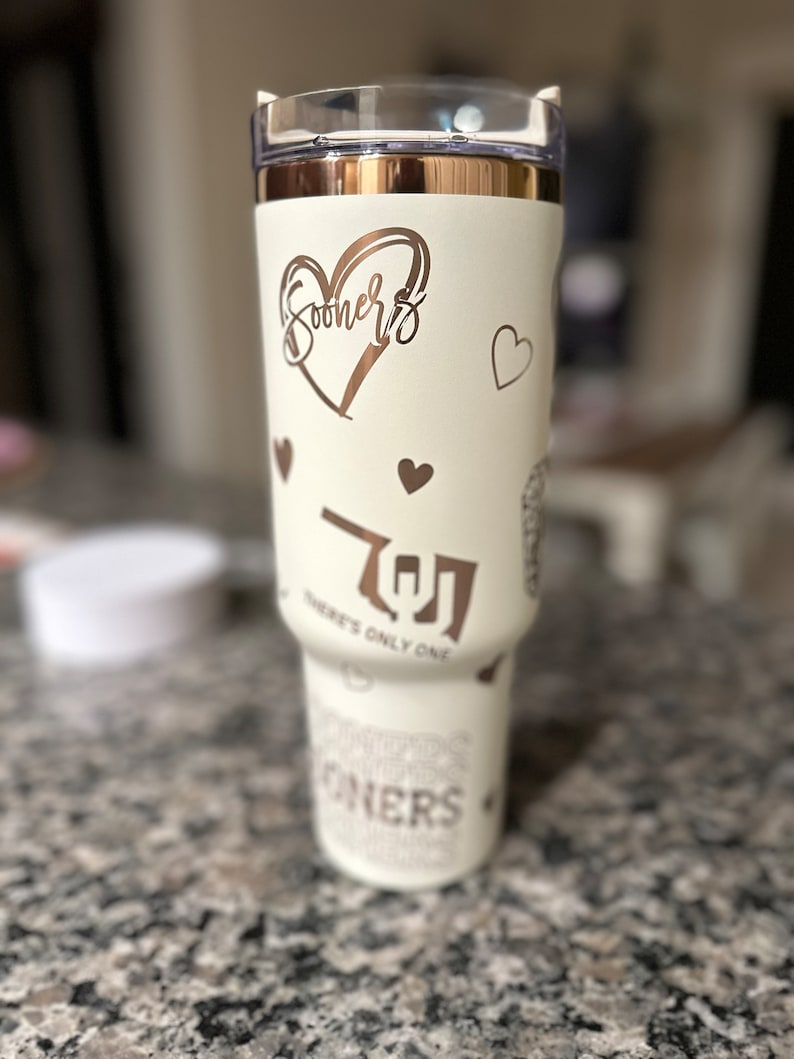 May include: White insulated tumbler with a rose gold lid and rose gold lettering. The tumbler features a heart with the word "Sooners" inside, a heart with the letter "O" inside, and the text "There's Only One".