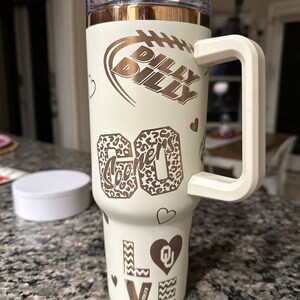 May include: White stainless steel tumbler with a gold lid and handle. The tumbler has a variety of designs including a football, hearts, and the words "Billy Billy" and "Sooners".