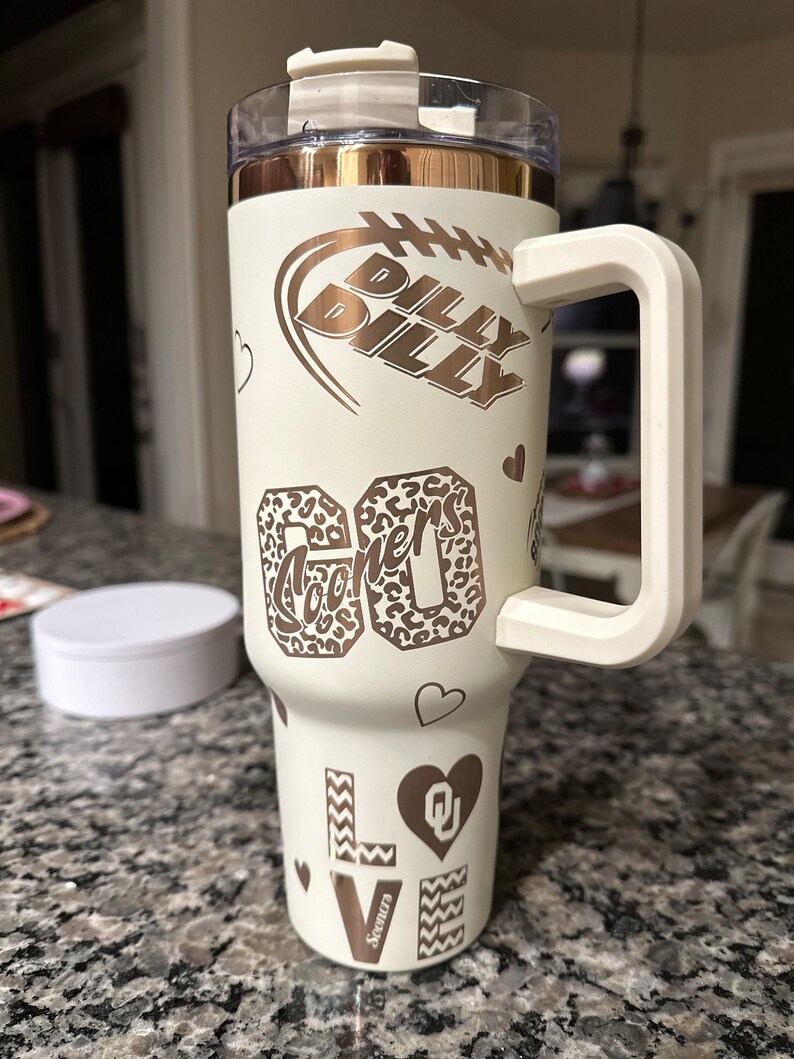 May include: White stainless steel tumbler with a gold lid and handle. The tumbler has a variety of designs including a football, hearts, and the words "Billy Billy" and "Sooners".