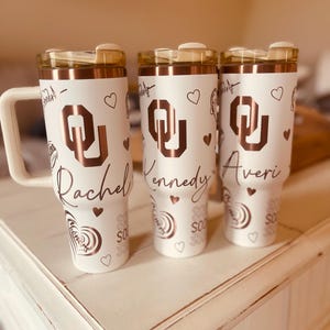 May include: Three white personalised tumblers with a rose gold design. The tumblers feature a university logo and the names Rachel, Kennedy, and Averi.