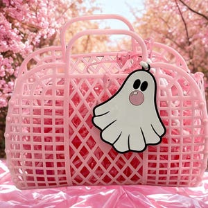 May include: A pink plastic basket with a diamond lattice design and two handles. A white ghost-shaped tag with black eyes and a pink bubble is attached to the handle. The basket is filled with pink material and sits on a pink surface.