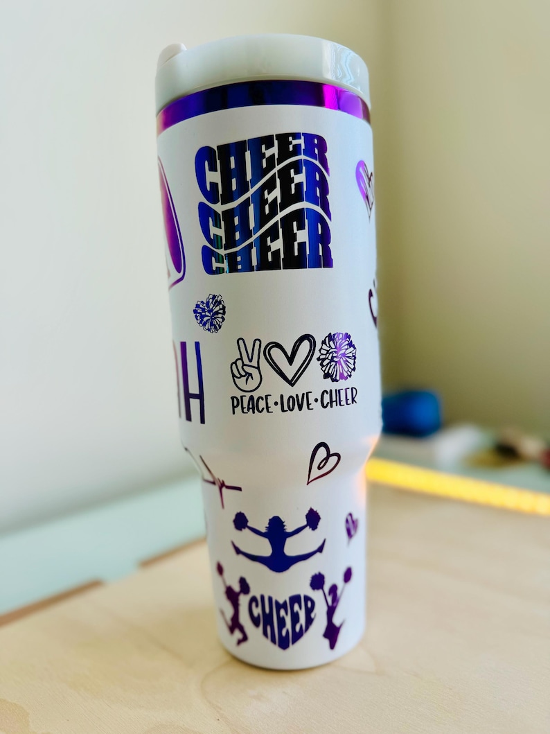 May include: White tumbler with a purple iridescent lid and purple cheerleading themed decals. The decals include the words "Cheer" three times, "Peace-Love-Cheer", and a silhouette of a cheerleader.