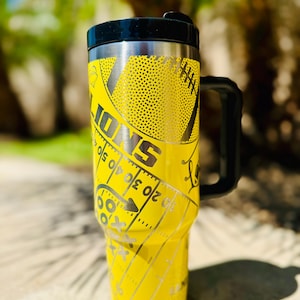 May include: Yellow stainless steel travel mug with a black lid and handle. The mug has a football design with the word "IONS" and a football field graphic.