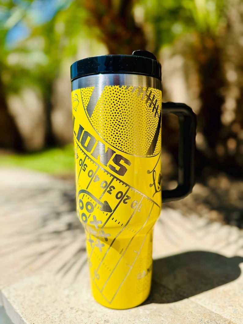 May include: Yellow stainless steel travel mug with a black lid and handle. The mug has a football design with the word "IONS" and a football field graphic.
