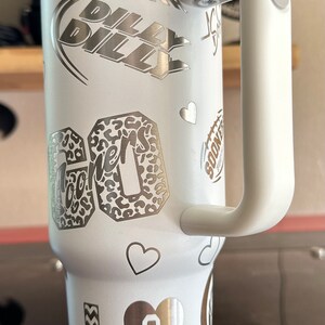 May include: White stainless steel travel mug with a black and white graphic of a football and the words "Billy Billy". The mug also has a graphic of a leopard print with the word "Sooners" and a heart with the letters "OU".