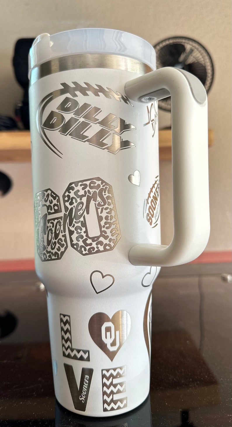 May include: White stainless steel travel mug with a black and white graphic of a football and the words "Billy Billy". The mug also has a graphic of a leopard print with the word "Sooners" and a heart with the letters "OU".