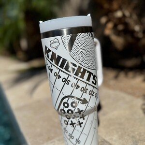 May include: White stainless steel travel mug with a black and white design featuring a football, the word "Knights", and a football field with yard lines.
