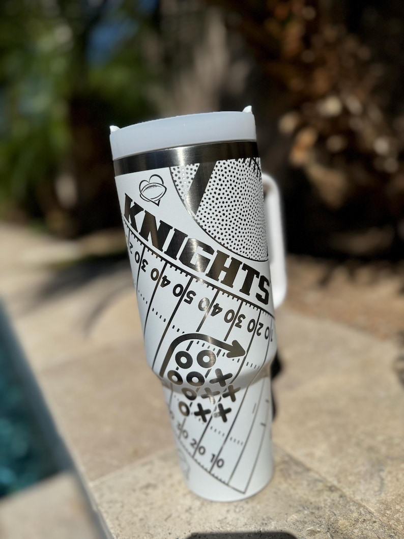May include: White stainless steel travel mug with a black and white design featuring a football, the word "Knights", and a football field with yard lines.