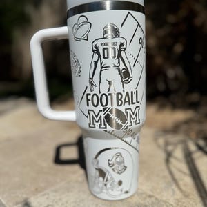 May include: White stainless steel travel mug with a black and white design featuring a football player, a football helmet, and the text "Football Mom".