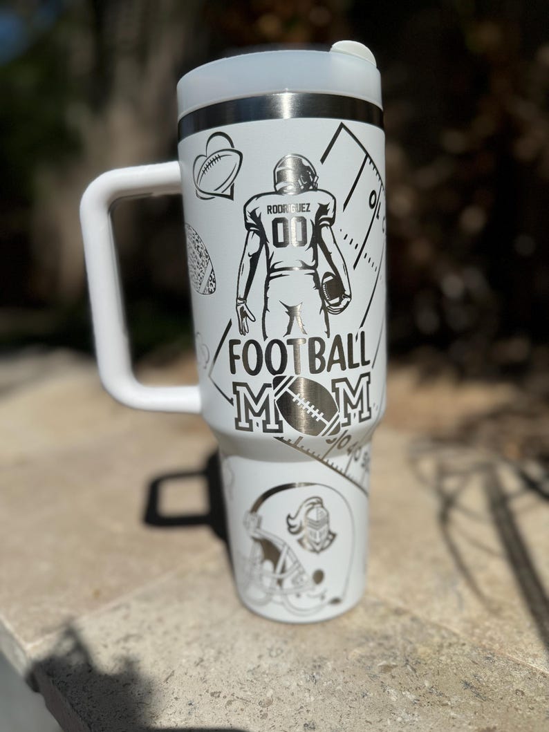 May include: White stainless steel travel mug with a black and white design featuring a football player, a football helmet, and the text "Football Mom".