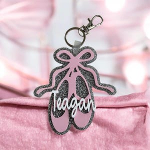 May include: A pink and glittery ballet slipper keyring with the name "Teagan" in white script. The keyring has a silver clasp and ring. The design includes a bow and ribbon detail, set against a soft pink background.